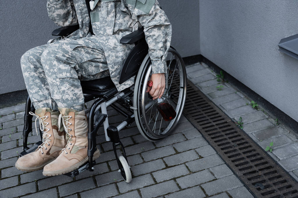bijgesneden beeld van een gehandicapte soldaat die in een rolstoel zit en buiten een fles whisky vasthoudt - Foto, afbeelding