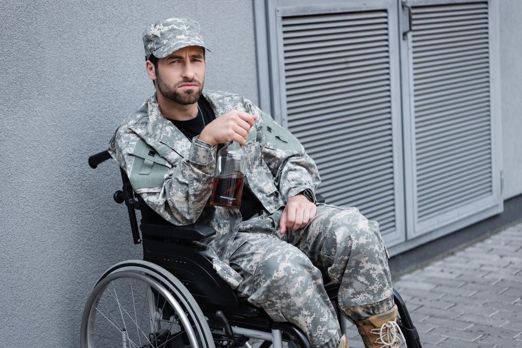καταθλιπτικός και ανάπηρος στρατιωτικός που κάθεται σε αναπηρική καρέκλα με μπουκάλι αλκοόλ - Φωτογραφία, εικόνα