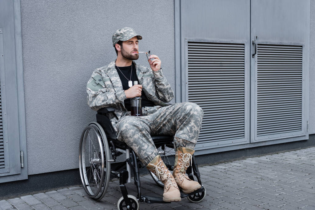 ветеран-инвалид в инвалидной коляске закуривает сигарету и держит бутылку алкоголя на открытом воздухе - Фото, изображение