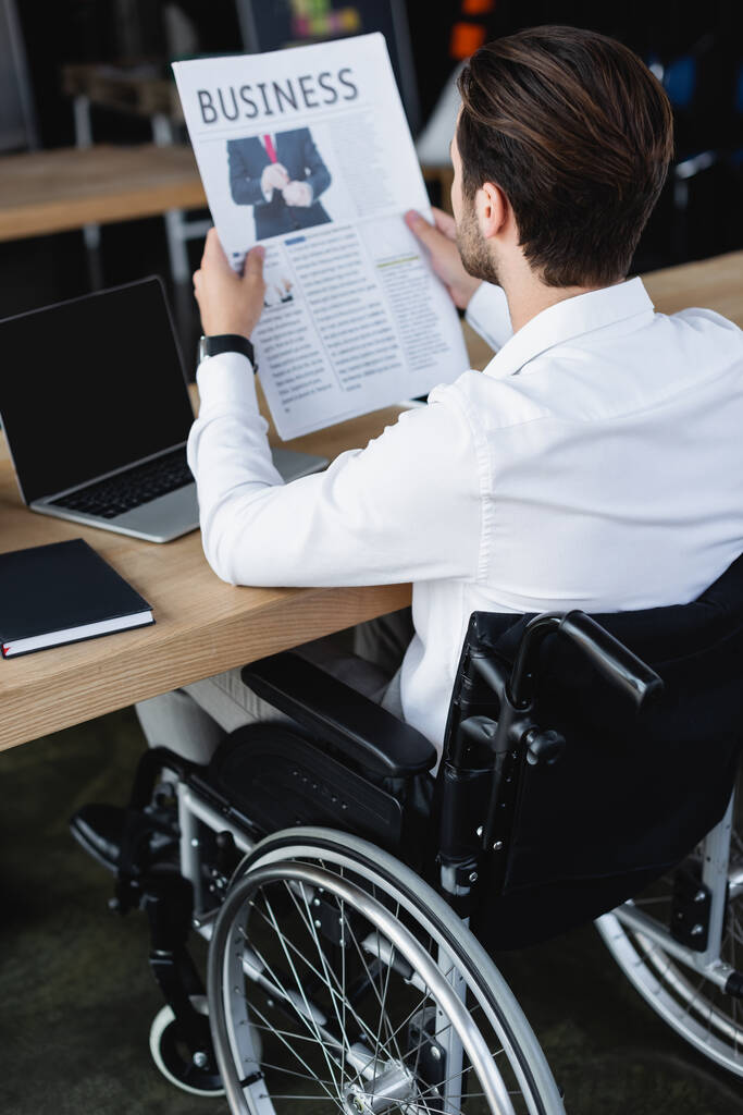 Tekerlekli sandalyedeki genç iş adamı boş ekranla dizüstü bilgisayar başında gazete okuyor. - Fotoğraf, Görsel