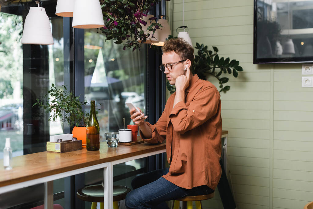 молодой человек в очках регулирует беспроводные наушники и использует смартфон в кафе  - Фото, изображение
