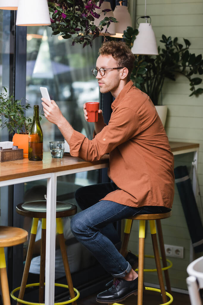 young man in glasses holding cup and using smartphone in cafe  - Photo, Image