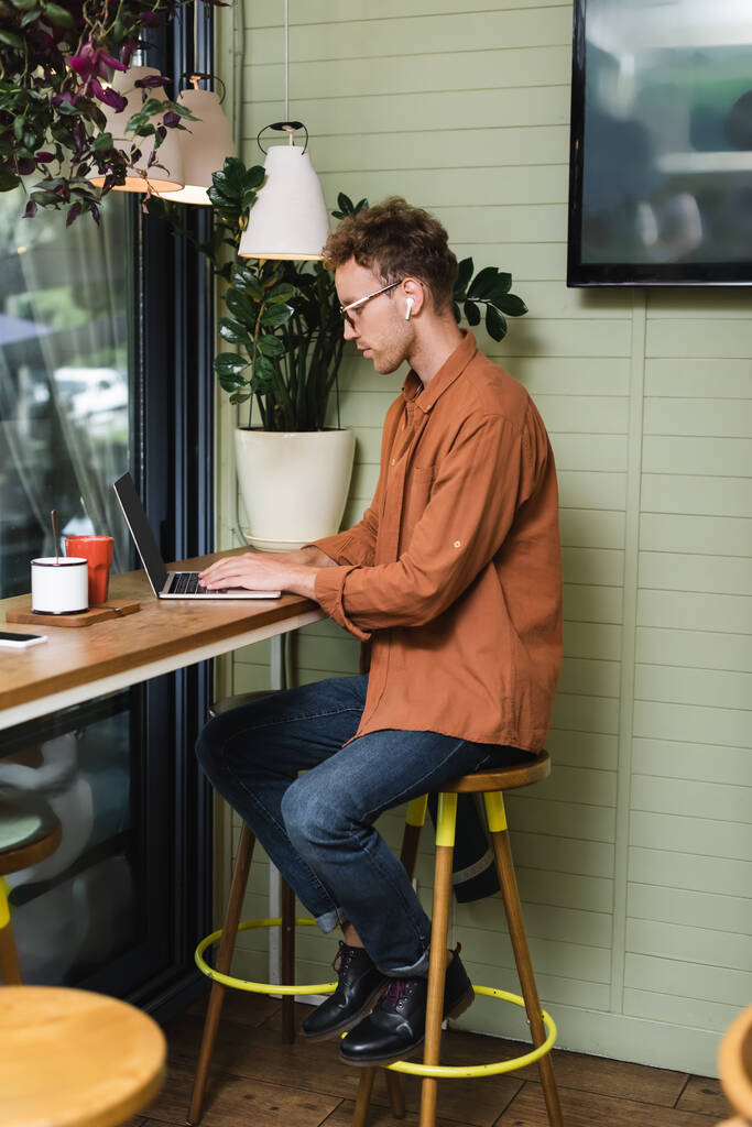 вид збоку молодого чоловіка в окулярах, що друкуються на ноутбуці в кафе
 - Фото, зображення