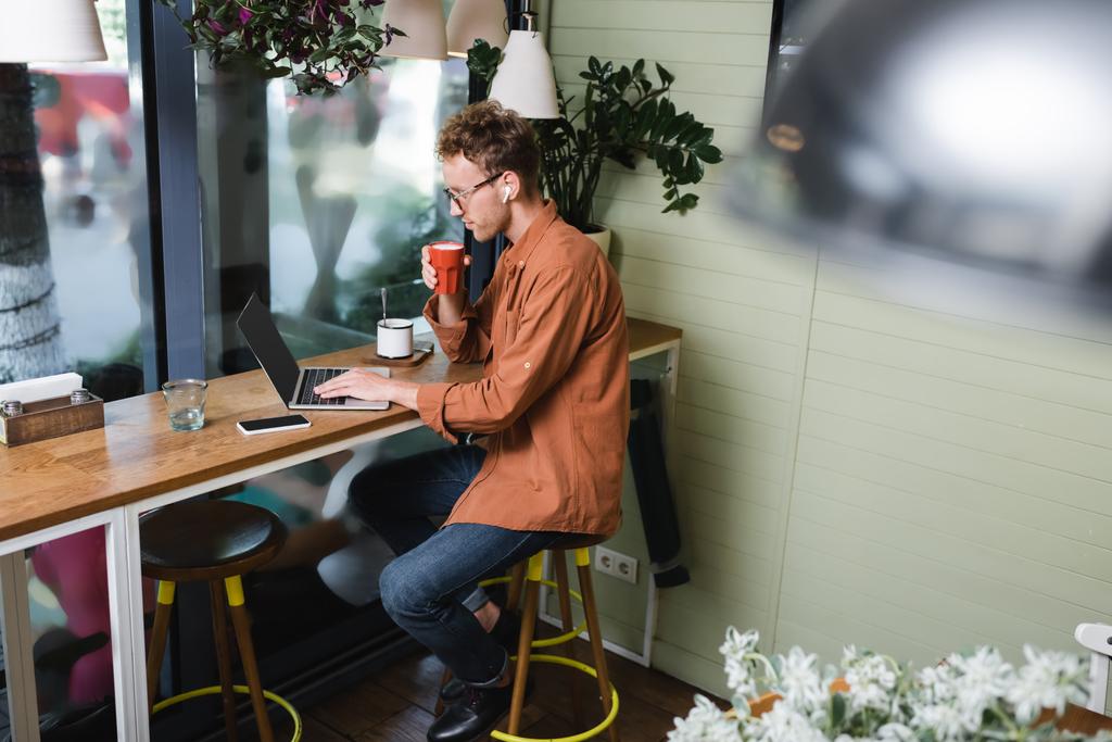 ελεύθερος επαγγελματίας σε ακουστικά κρατώντας κύπελλο και χρησιμοποιώντας φορητό υπολογιστή κοντά στο κινητό τηλέφωνο στο τραπέζι στο καφέ  - Φωτογραφία, εικόνα