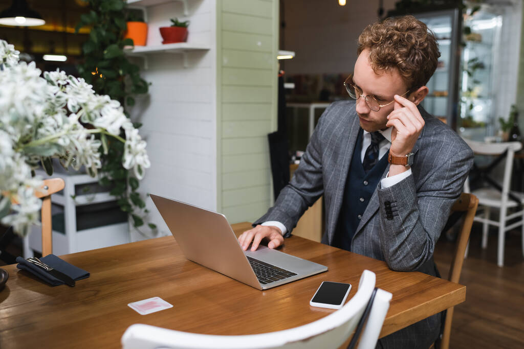 homem de negócios encaracolado em terno ajustando óculos ao usar laptop no café - Foto, Imagem