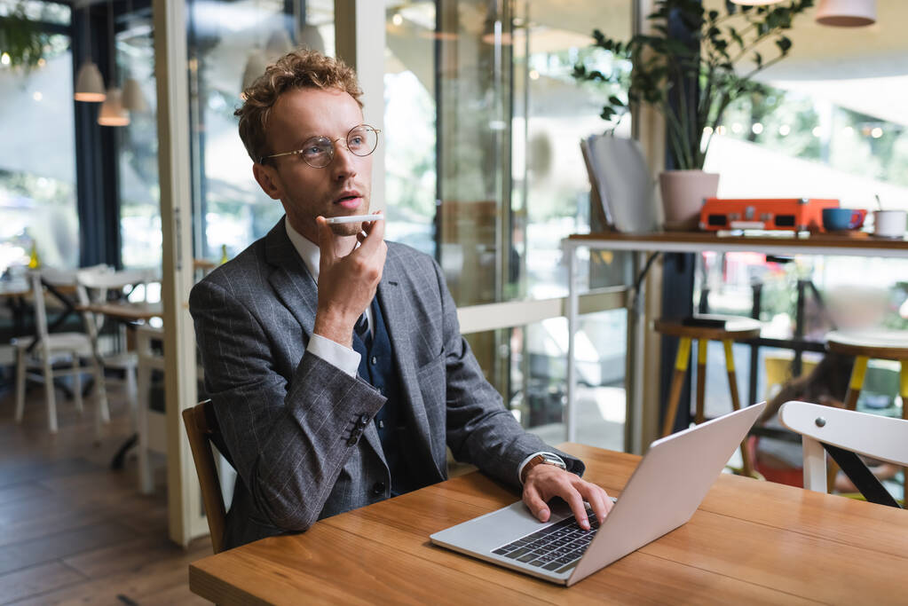 homem de negócios encaracolado em óculos gravando mensagem de voz no telefone celular perto do laptop  - Foto, Imagem