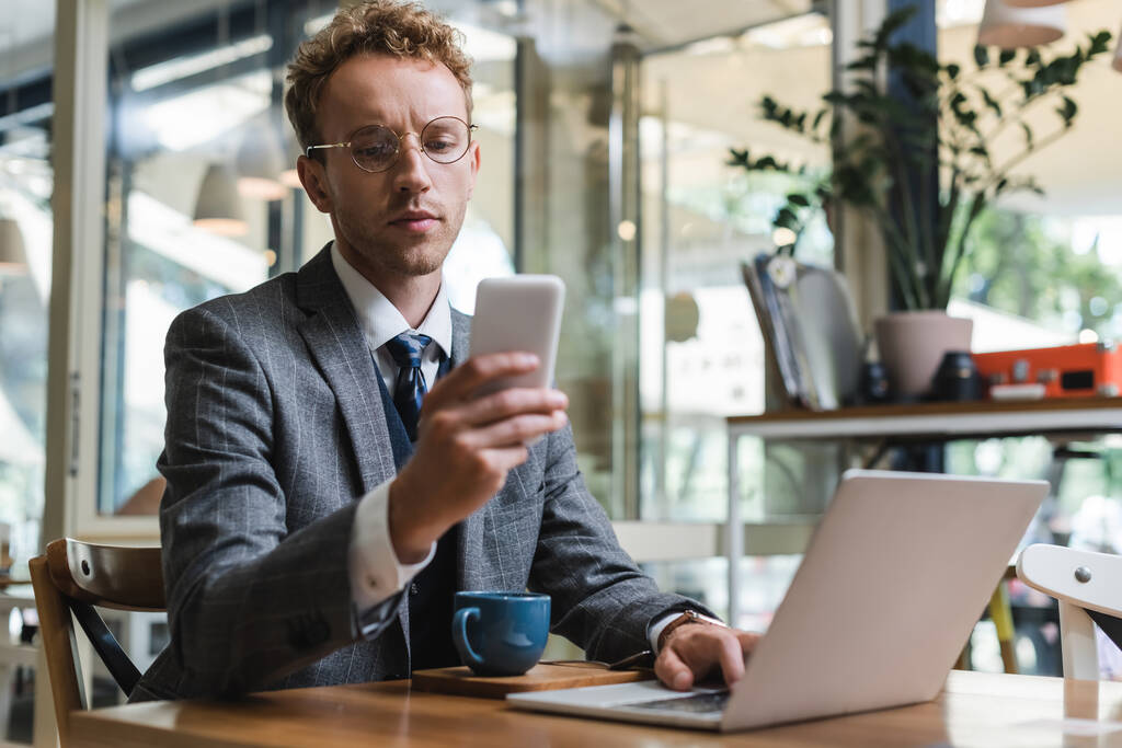 кудрявый бизнесмен в очках с помощью мобильного телефона возле ноутбука и чашки с кофе в кафе - Фото, изображение