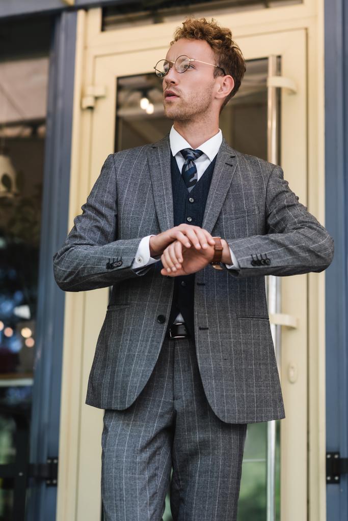 кучерявий бізнесмен в стильному костюмі регулювання наручних годинників біля входу в кафе
 - Фото, зображення