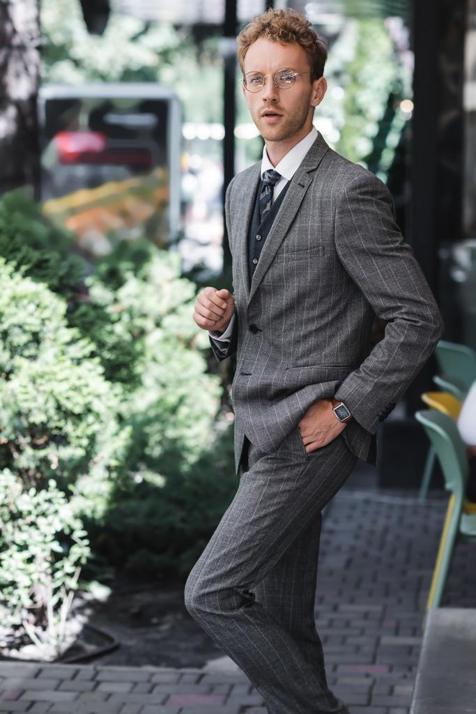 молодой бизнесмен в очках и стильном костюме, стоящий с рукой в кармане возле входа в кафе - Фото, изображение