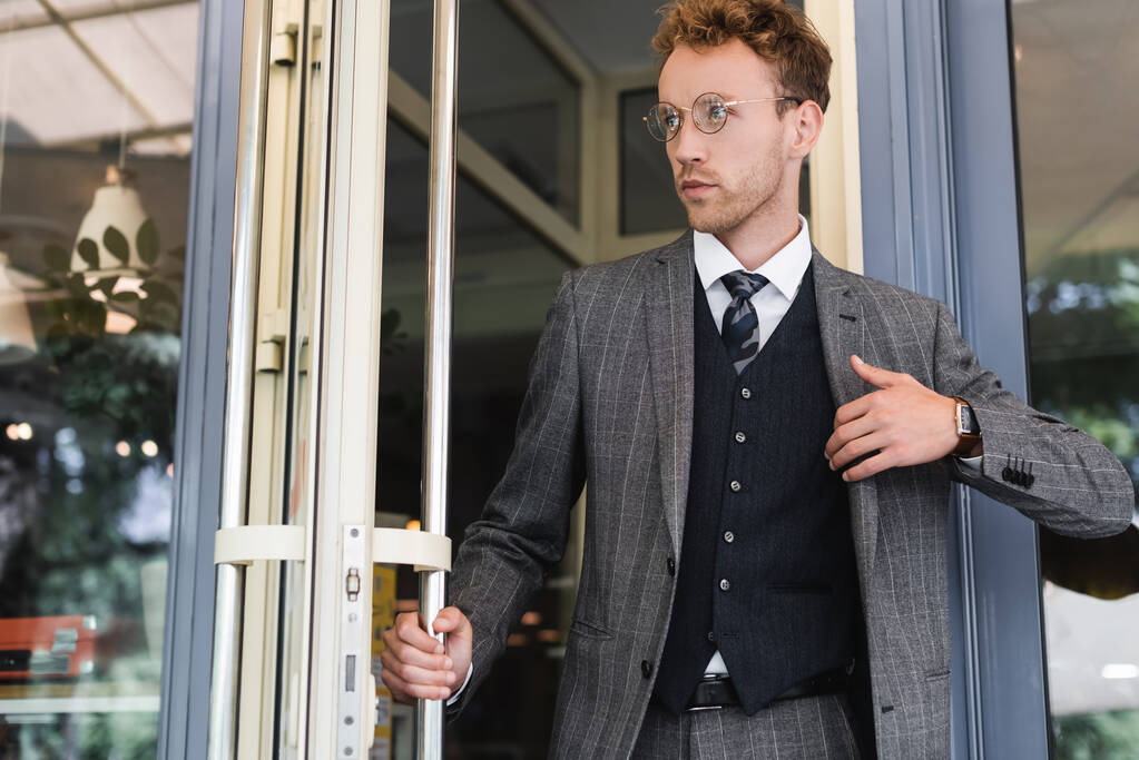junger, lockiger Geschäftsmann mit Brille öffnet Tür beim Verlassen des Cafés  - Foto, Bild