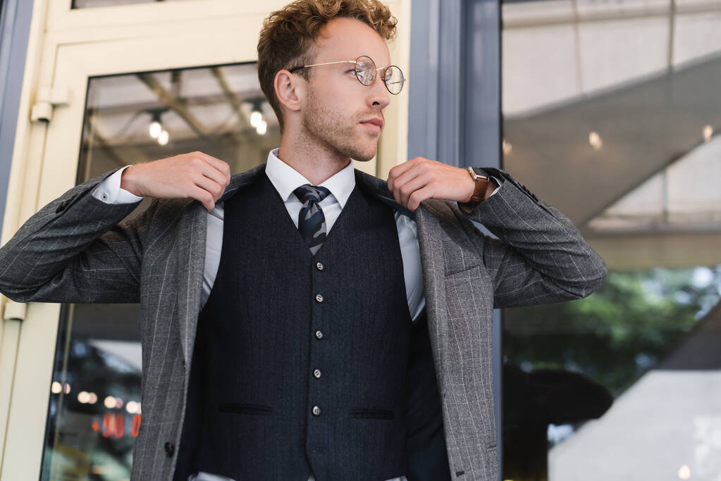 σγουρός επιχειρηματίας με γυαλιά και κομψό κοστούμι φορώντας σακάκι κοντά στην είσοδο του καφέ - Φωτογραφία, εικόνα