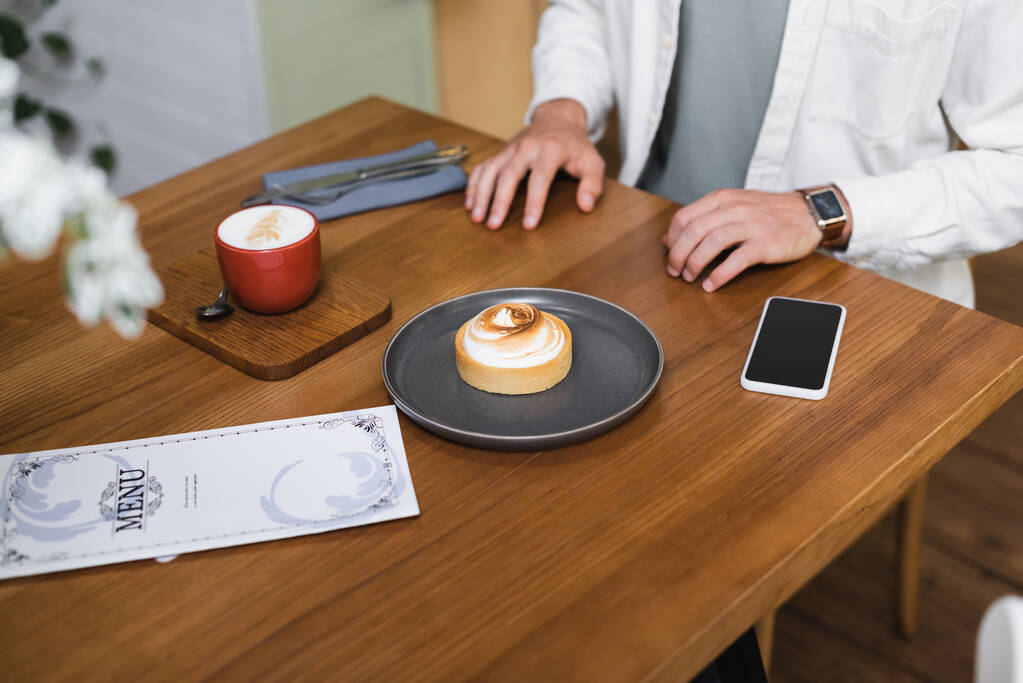vágott kilátás ember közelében citromos torta, cappuccino, menü és okostelefon üres képernyő az asztalon  - Fotó, kép