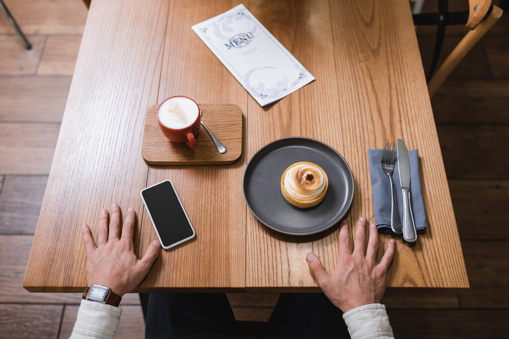 レモンタルト近くの男の高い角度ビュー,テーブルの上に空白の画面でスマートフォンの近くのカプチーノとメニュー  - 写真・画像