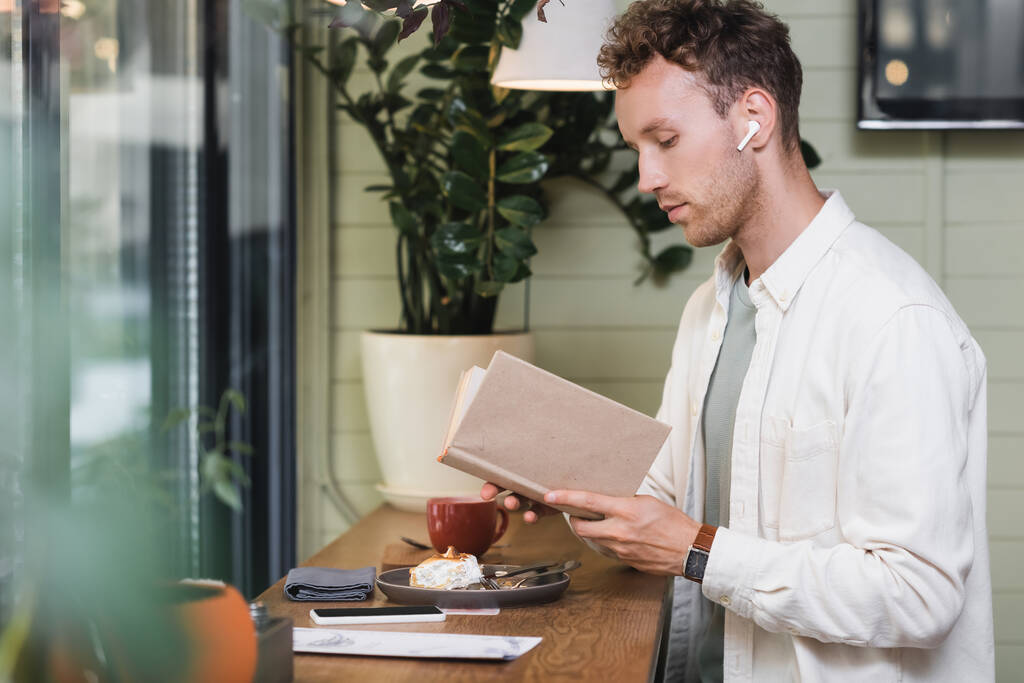 göndör férfi vezeték nélküli fülhallgató olvasás könyv kávézóban  - Fotó, kép