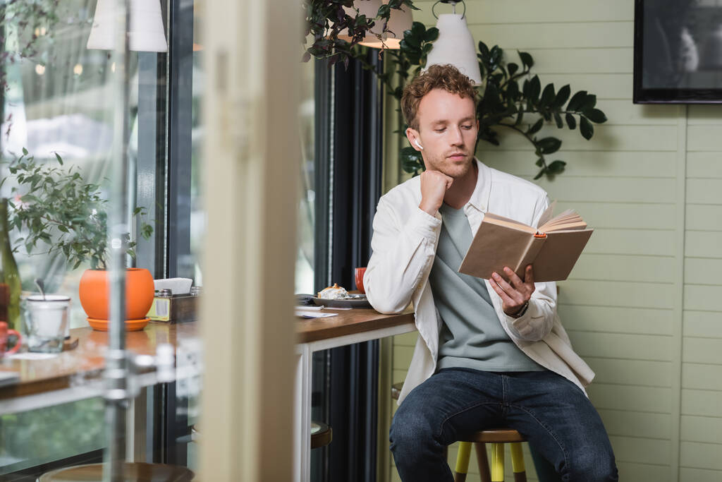 Kablosuz kulaklıklarda odaklanmış adam kafede kitap okuyor. - Fotoğraf, Görsel