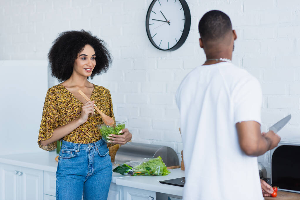 Positiivinen afrikkalainen amerikkalainen nainen sekoittamalla salaattia lähellä hämärtynyt poikaystävä veitsellä keittiössä  - Valokuva, kuva