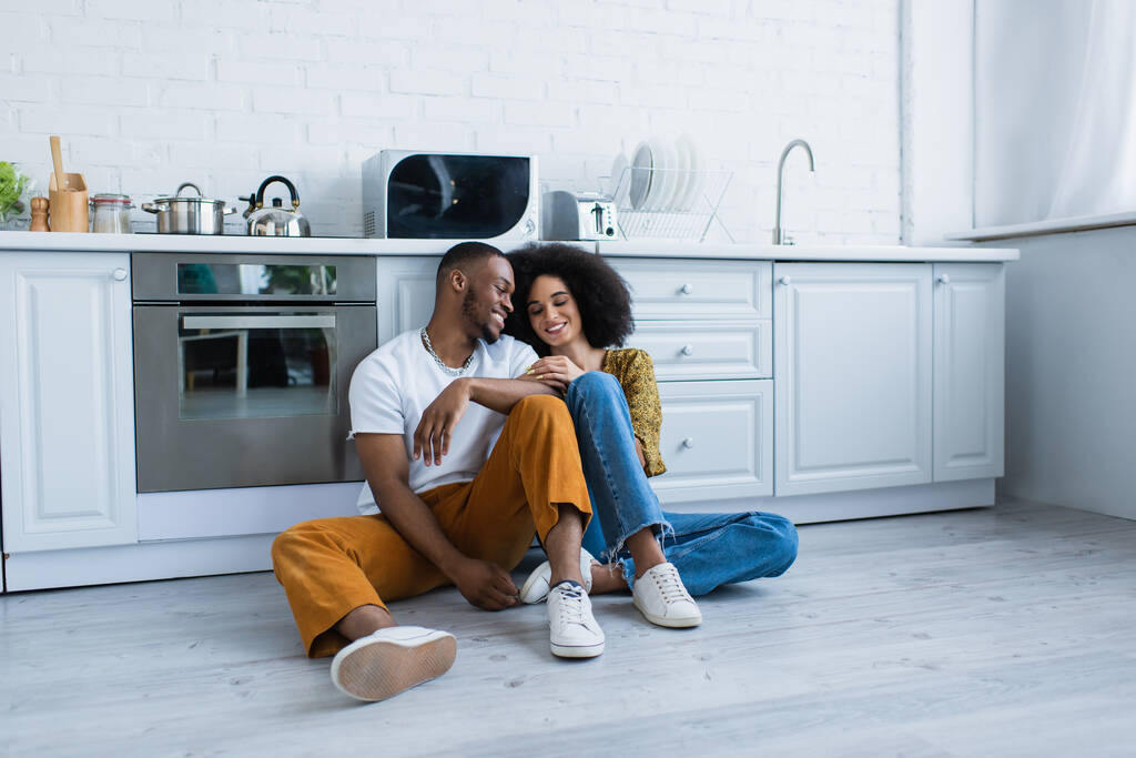Africano americano homem sentado perto sorrindo namorada no chão na cozinha  - Foto, Imagem