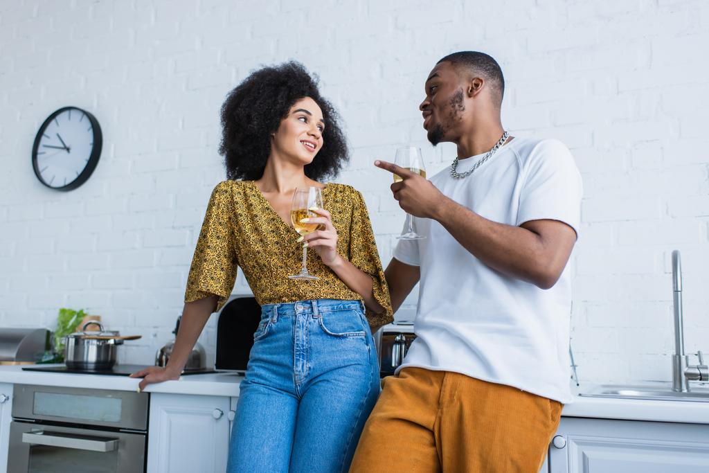 African american couple with glasses of wine talking near worktop in kitchen  - Photo, Image