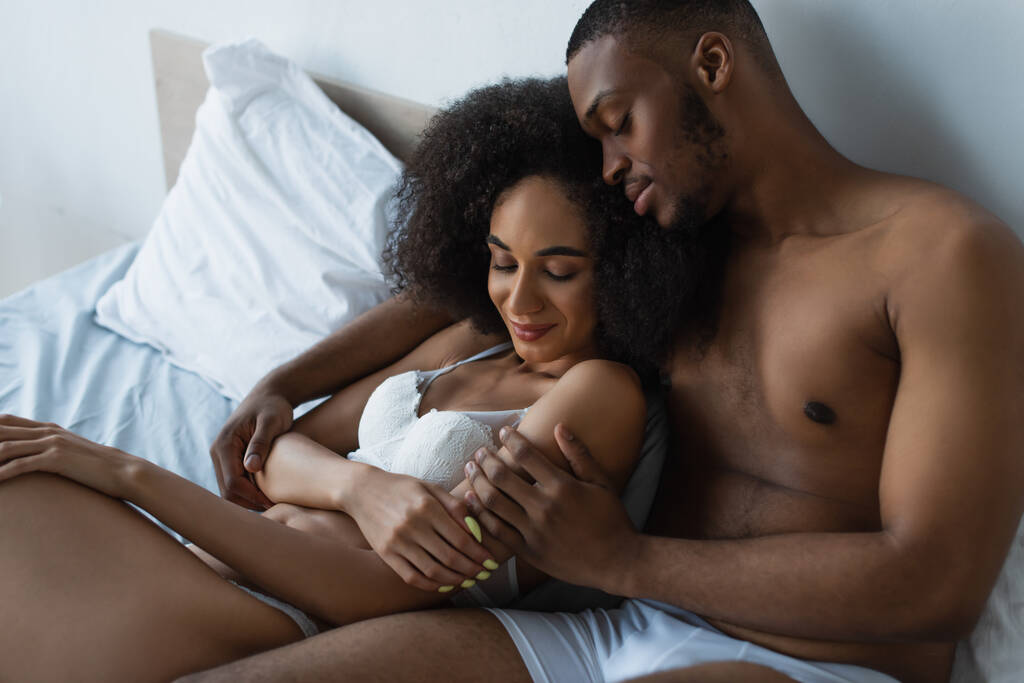 Africano americano hombre con los ojos cerrados abrazando novia en sujetador en la cama  - Foto, imagen