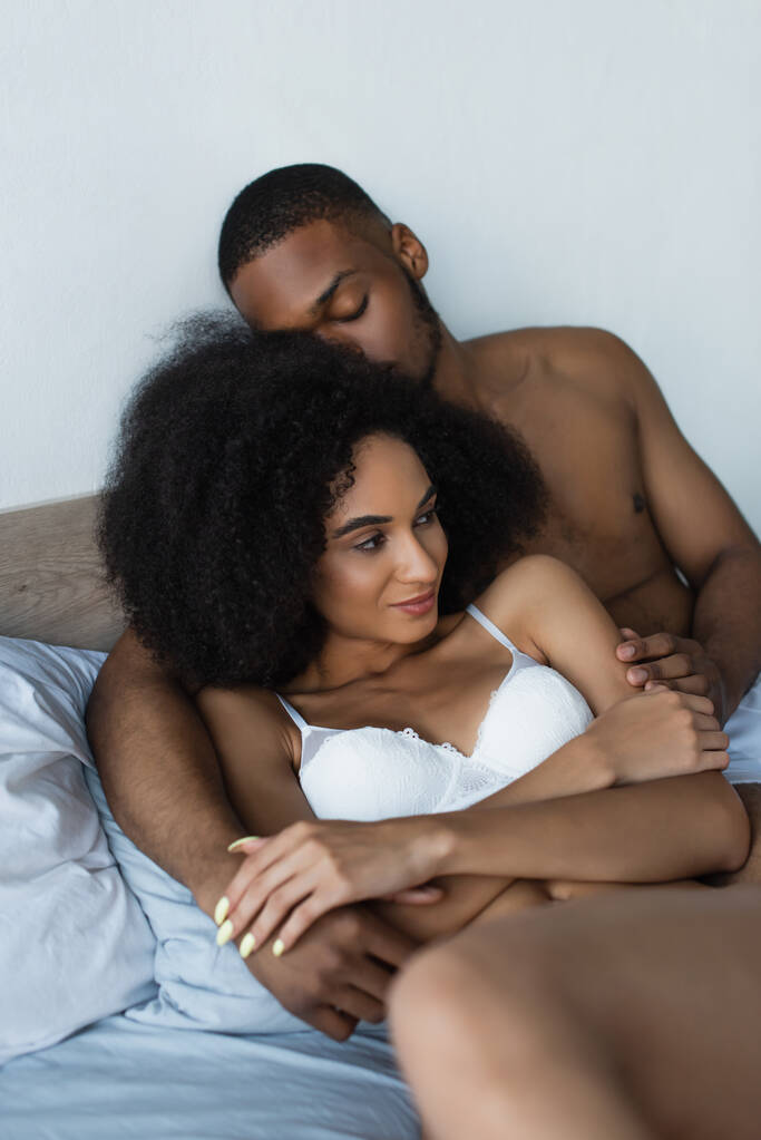 Junger afrikanisch-amerikanischer Mann berührt sinnliche Frau im BH auf Bett  - Foto, Bild