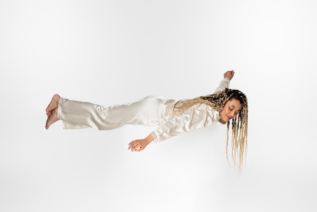 barefoot african american woman in silk pajamas sleeping in levitation isolated on white - Photo, Image