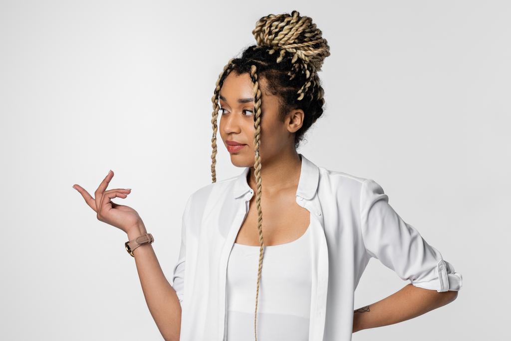 joven mujer de negocios afroamericana con rastas mirando hacia otro lado y señalando con el dedo aislado en blanco - Foto, Imagen