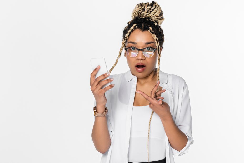 mulher de negócios afro-americana surpreendida apontando para smartphone enquanto olha para a câmera isolada no branco - Foto, Imagem