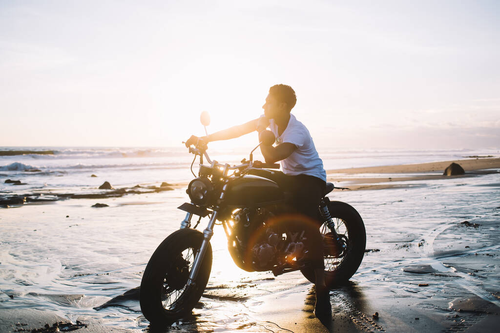 Side view of adult biker in casual clothes touching chin and pondering while sitting on motorcycle and looking away on wet ocean sand beach during sunset on background of clear sky and waves - Photo, Image