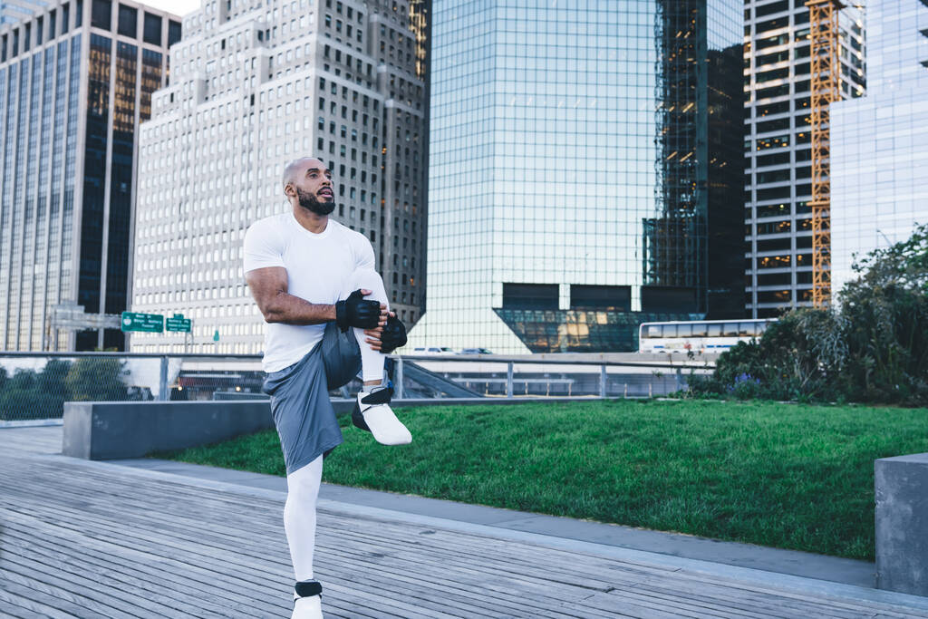 Gut gebauter schwarzer Sportler, der morgens zielstrebig auf dem hölzernen Bürgersteig an der Stadtstraße Übungen für die Beine praktiziert - Foto, Bild