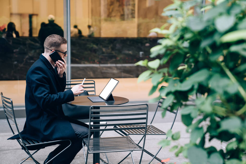 Zijaanzicht van toonbare en zelfverzekerde zakenman in strikte pak zitten buiten voor het raam en praten op de smartphone, terwijl het kijken naar lege scherm van de tablet - Foto, afbeelding