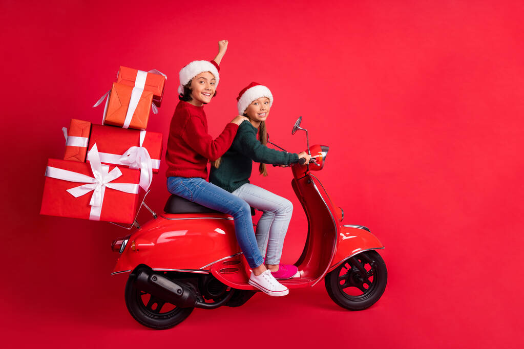 Φωτογραφία από αστεία όμορφα σχολιαρόπαιδα ντυμένοι πλεκτό πουλόβερ ιππασίας μοτοποδήλατο μεταφέρουν δώρα απομονωμένο φόντο κόκκινο χρώμα - Φωτογραφία, εικόνα