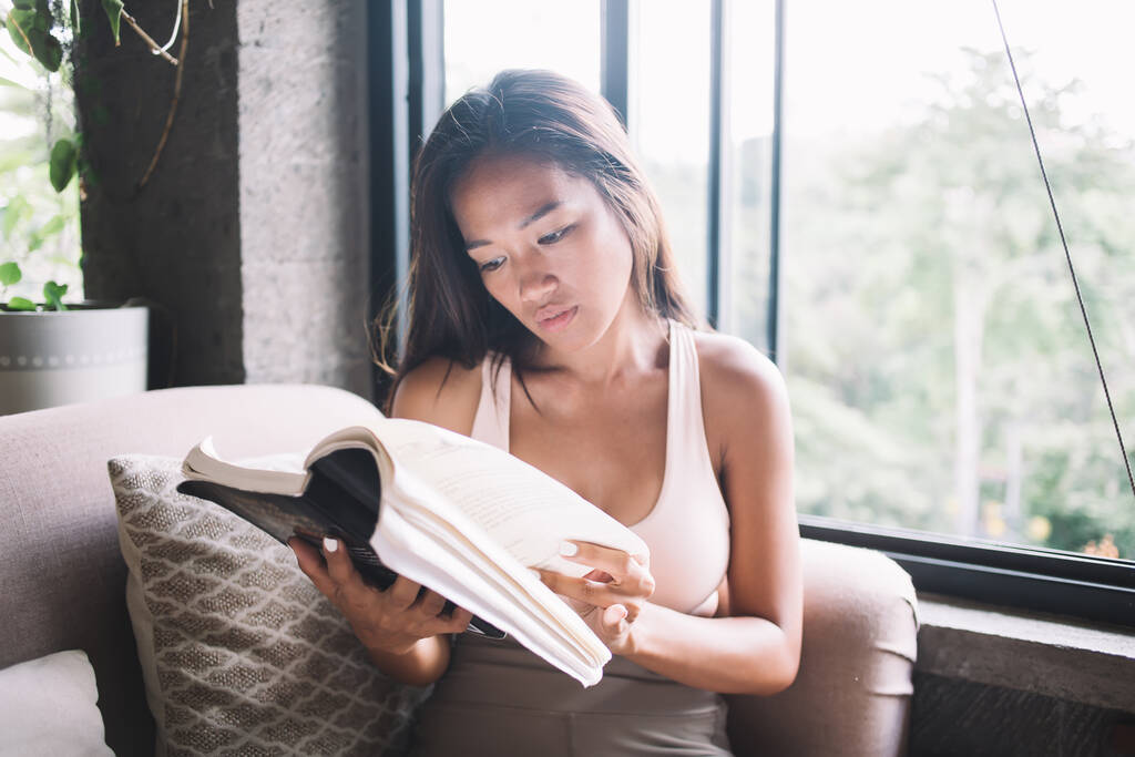 Giovane focalizzata etnica femminile in reggiseno sportivo flipping pagine di libro durante la lettura sul divano contro finestra in appartamento luce - Foto, immagini