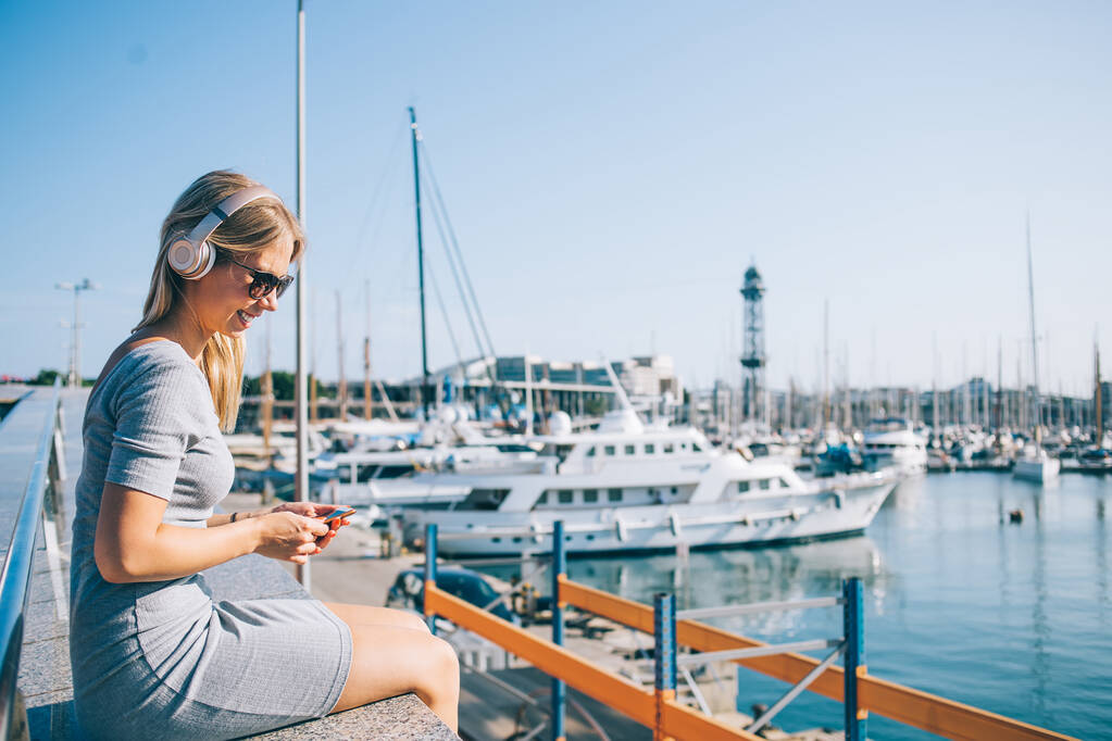 Side view of happy young lady in headphones and sunglasses smiling and browsing smartphone while sitting and listening to music in port against cloudless sky in Barcelona City - Photo, Image
