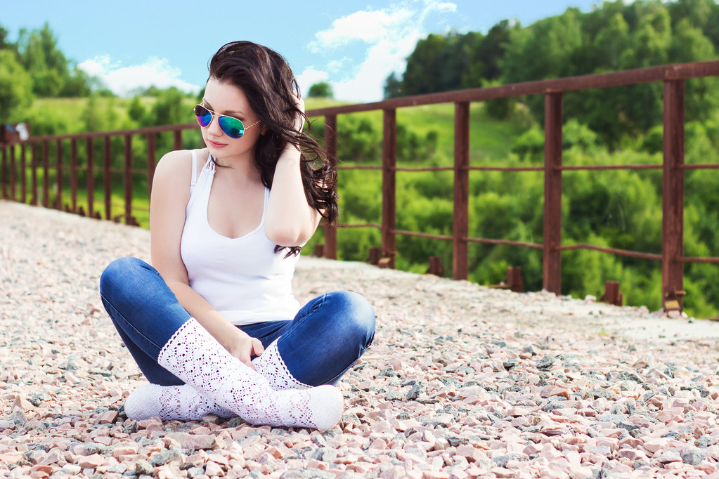 Menina bonita com cabelo preto longo em um dia ensolarado de verão sentado na ponte em óculos de sol
 - Foto, Imagem