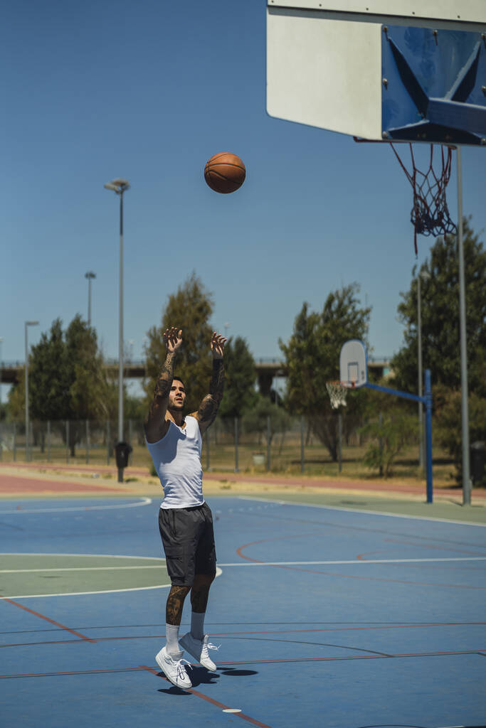 Um tiro vertical de um cara caucasiano com tatuagens legais jogando a bola de basquete para o aro - Foto, Imagem