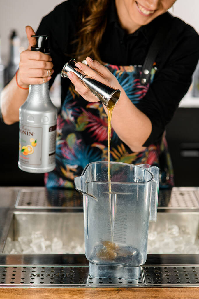 UKRAINE, KIEW - 12. MÄRZ 2021: Barkeeperin hält Stahlvorrichtung und gießt Sirup daraus in Mixerglas. Flasche Marke Monin in der Hand - Foto, Bild