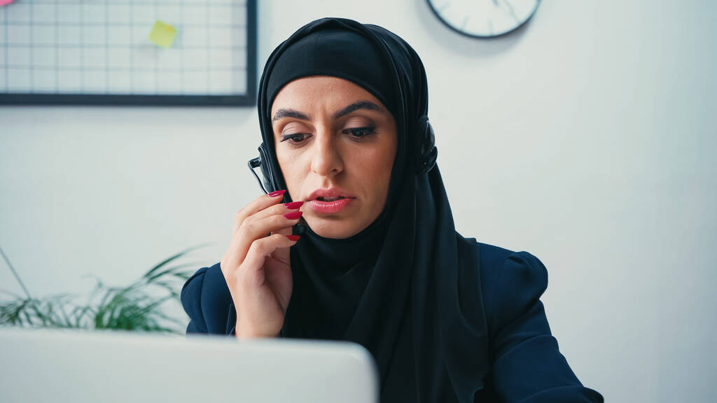 Mujer joven musulmana en hijab con auriculares cerca de la computadora portátil borrosa en el centro de llamadas  - Foto, Imagen