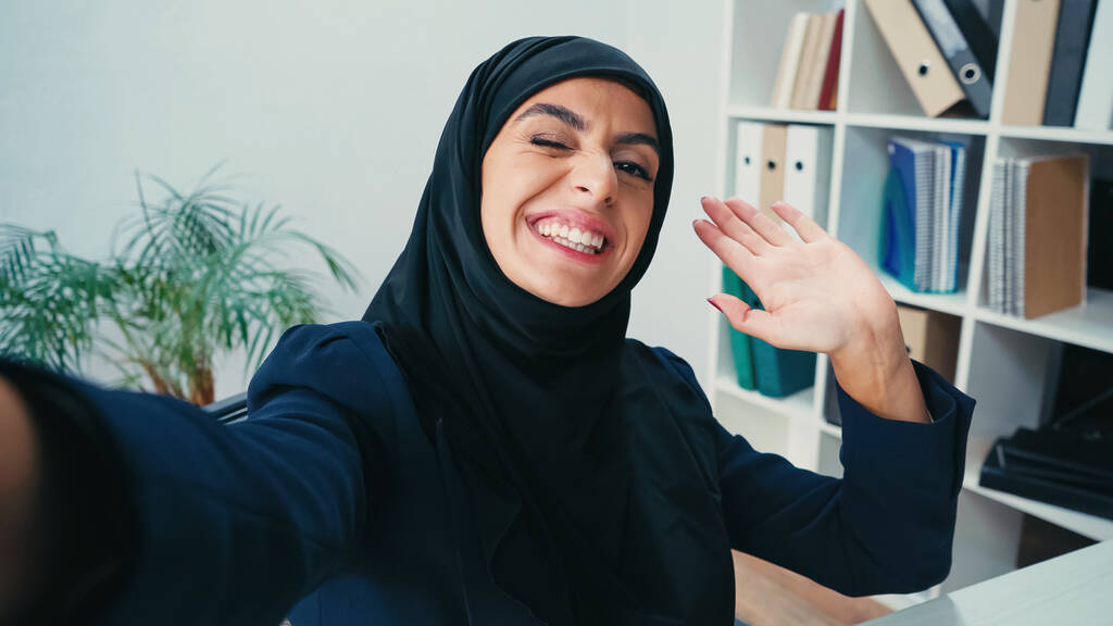 Улыбающаяся мусульманка-бизнесмен машет рукой, делая селфи  - Фото, изображение