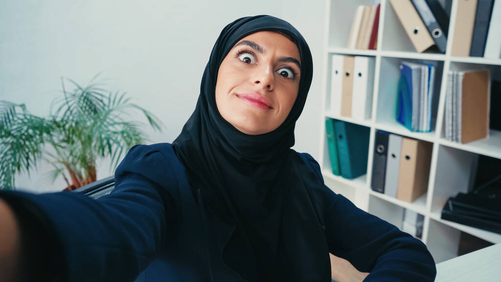 sonriente joven musulmana mujer de negocios haciendo muecas mientras toma selfie  - Foto, Imagen