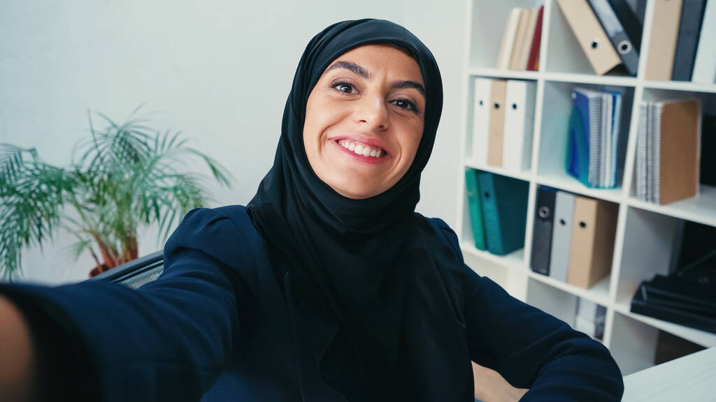 cheerful muslim businesswoman looking at camera while taking selfie  - Photo, Image