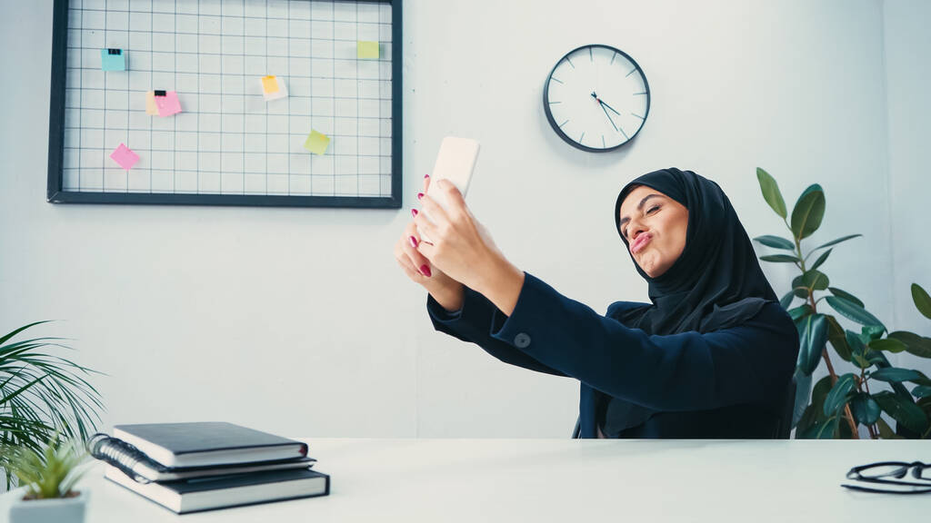 muzułmanka bizneswoman dąsając usta i biorąc selfie na smartfonie w biurze  - Zdjęcie, obraz
