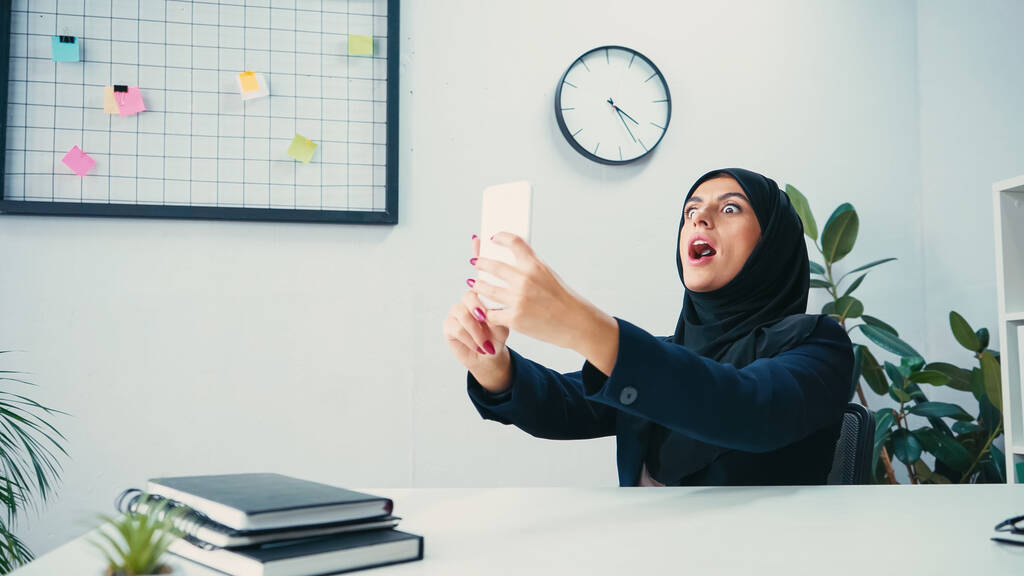 Μουσουλμάνα επιχειρηματίας με ανοιχτό στόμα που βγάζει selfie στο smartphone στο γραφείο  - Φωτογραφία, εικόνα