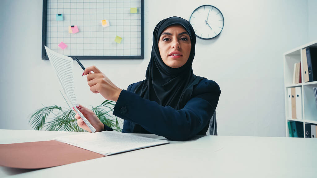 Мусульманская женщина в хиджабе указывает ручкой на документ в современном офисе  - Фото, изображение