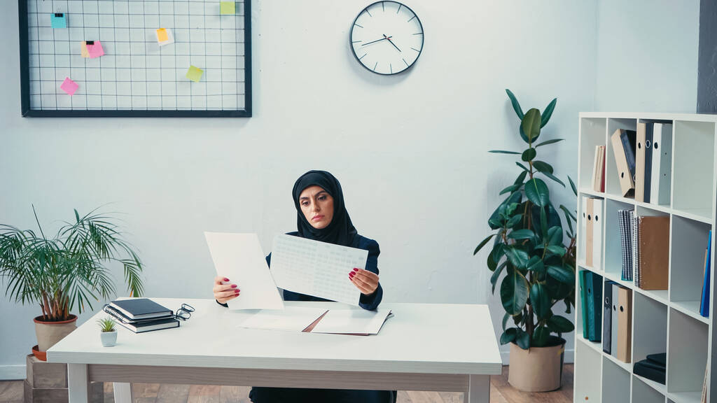 Junge muslimische Frau im Hidschab schaut sich Dokumente an und sitzt am Schreibtisch im Büro - Foto, Bild