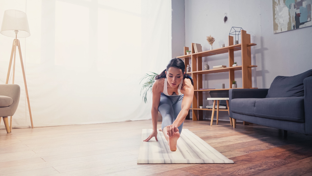 Подходит женщина, растягивающая ногу на фитнес-коврике в гостиной  - Фото, изображение