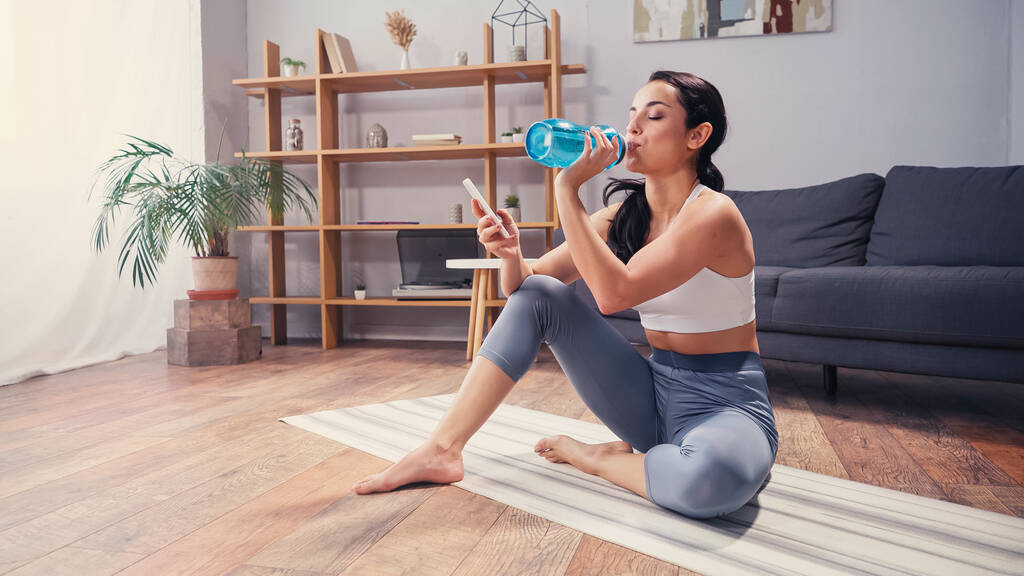 Desportista água potável e usando celular no tapete de fitness na sala de estar  - Foto, Imagem