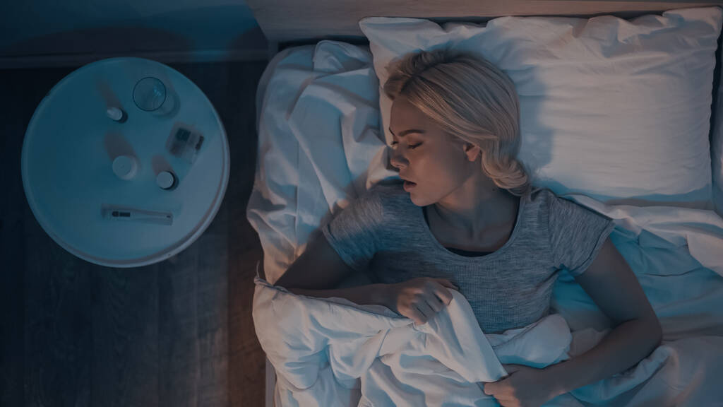 Vista superior de la mujer joven durmiendo cerca del termómetro y pastillas en la mesita de noche  - Foto, Imagen