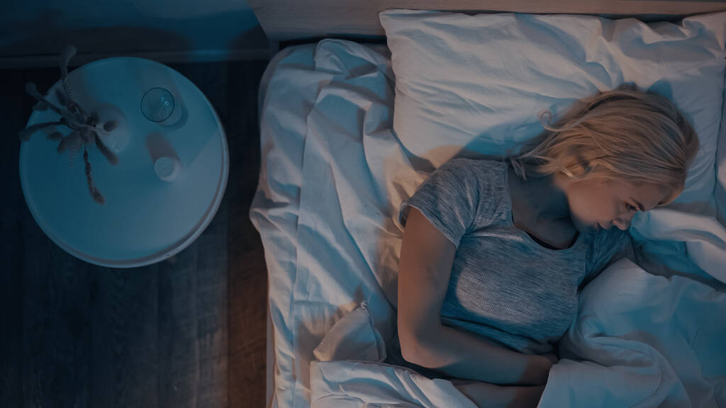 Horní pohled na mladou ženu ležící na posteli v blízkosti pilulek a vody na nočním stolku  - Fotografie, Obrázek
