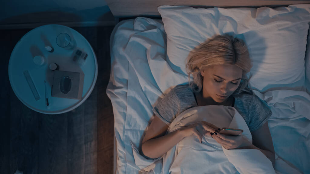 Draufsicht auf junge Frau mit Smartphone in der Nähe von Pillen und elektronischem Thermometer auf dem Bett  - Foto, Bild