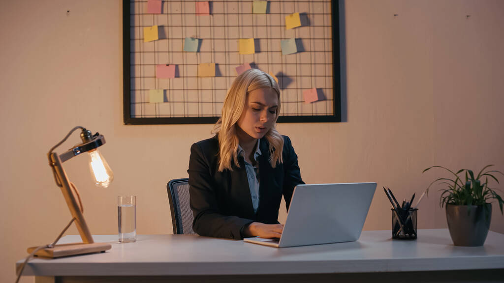 Бізнес-леді, використовуючи ноутбук біля лампи в офісі ввечері
  - Фото, зображення